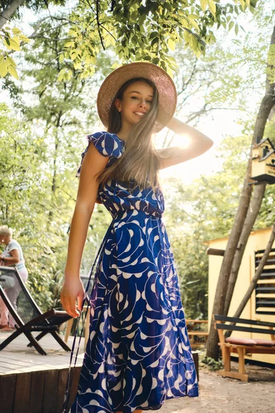 Młoda Ładna uśmiechnięta kobieta w niebieskiej sukni i kapelusz szczęśliwie patrząc w aparacie w pięknym parku miejskim — Zdjęcie stockowe