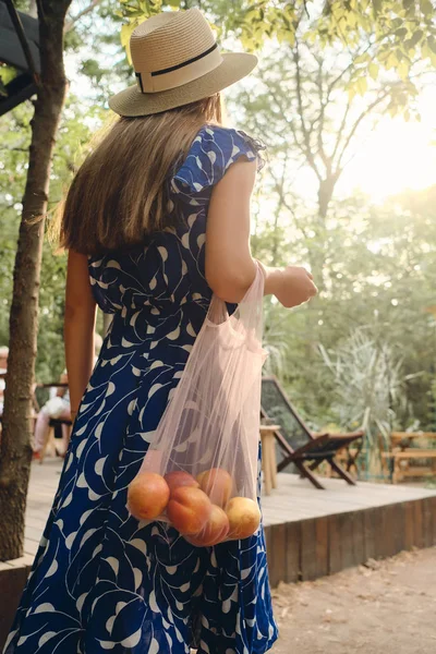 Wanita berambut coklat muda dengan gaun biru dan topi dengan buah persik di tas eko berdiri dari belakang di taman kota yang indah — Stok Foto