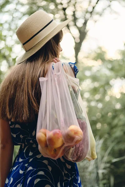Jonge bruin harige vrouw in blauwe jurk en hoed met Eco tassen met groenten en fruit op schouder staande van terug in prachtige stadspark — Stockfoto