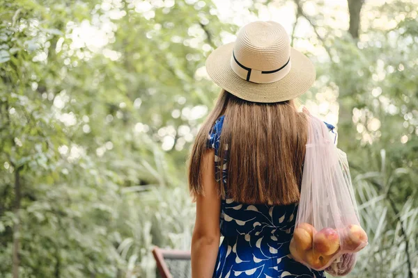 Jeune femme aux cheveux bruns en robe et chapeau tenant des sacs écologiques avec des fruits et légumes sur l'épaule debout de dos dans un beau parc — Photo