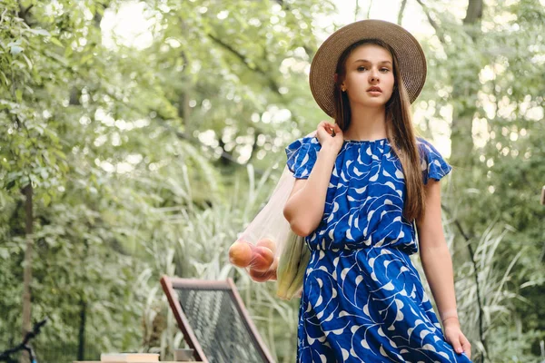 Joven hermosa mujer de cabello castaño en vestido azul y sombrero sosteniendo bolsas ecológicas con frutas y verduras en el hombro cuidadosamente mirando en la cámara en el parque —  Fotos de Stock