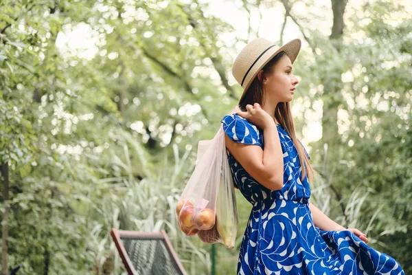 Красивая каштановые волосы девушка в синем платье и шляпа держа эко-мешки с фруктами и овощами на плече мечтательно глядя в сторону в красивом парке — стоковое фото