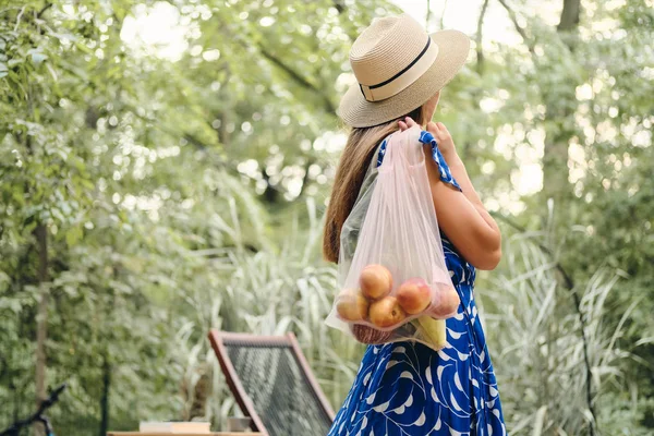 Jeune jolie femme aux cheveux bruns en robe et chapeau tenant des sacs écologiques avec des fruits et légumes sur l'épaule debout de dos dans un beau parc — Photo