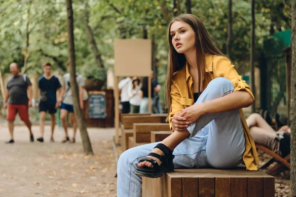 Joven atractiva chica adolescente de cabello castaño con camisa amarilla y jeans cuidadosamente mirando a un lado esperando a alguien en el banco de madera en el parque de la ciudad — Foto de Stock