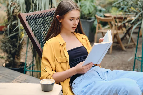 Jovem atraente adolescente de cabelos castanhos em camisa amarela e jeans livro de leitura sonhadora com café na cadeira de madeira deck no parque da cidade — Fotografia de Stock