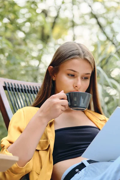 Wanita muda berambut cokelat yang menarik dengan kemeja kuning dan buku bacaan kelas atas dengan kopi di kursi dek kayu di taman kota — Stok Foto