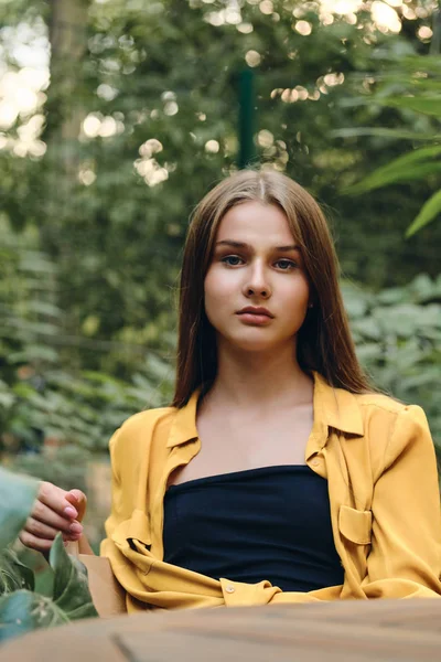 Jovem linda mulher de cabelos castanhos em camisa amarela e top seriamente olhando na câmera sentada no parque da cidade — Fotografia de Stock