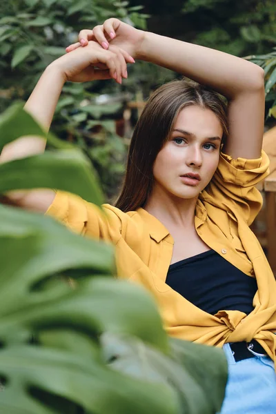 Young attractive brown haired woman in yellow shirt and top holding hand above head thoughtfully looking in camera around green leaves in city park — Stock Photo, Image