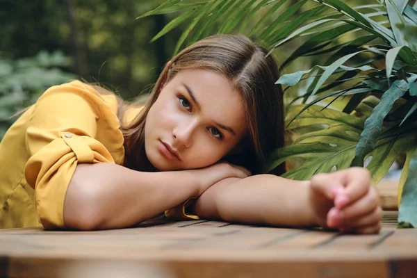 Young serious brown haired woman in yellow shirt lying on hand thoughtfully looking in camera around green leaves in park — Stock Photo, Image