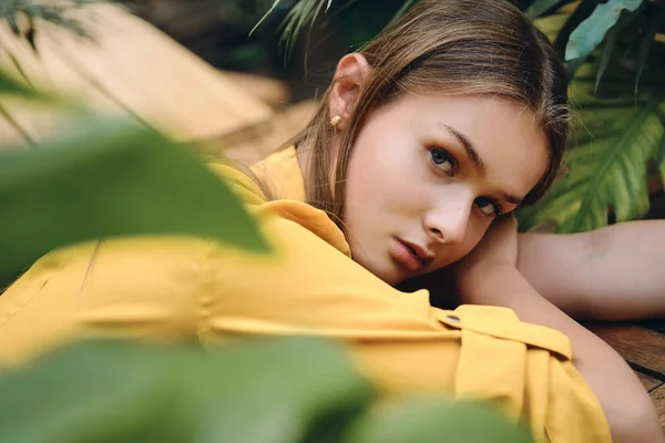 Jovem mulher de cabelos castanhos pensativo em camisa amarela deitado na mão olhando atenciosamente na câmera em torno de folhas verdes no parque — Fotografia de Stock