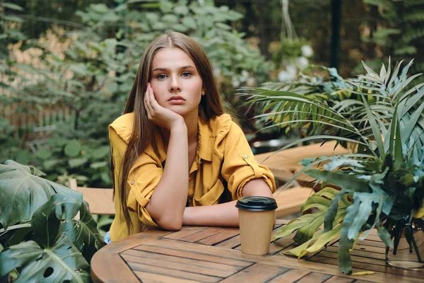 Jovem mulher séria em camisa amarela tristemente olhando na câmera sentada com xícara de café para ir no belo parque da cidade — Fotografia de Stock