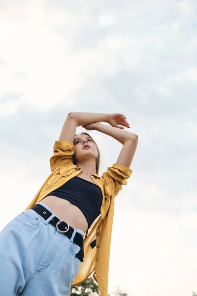 Νέοι ελκυστική γυναίκα με κίτρινο πουκάμισο και τζιν ονειρικά κρατώντας τα χέρια πάνω από το κεφάλι με ουρανό στο παρασκήνιο — Φωτογραφία Αρχείου