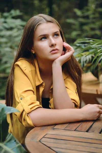 Young pensive brown haired woman in yellow shirt leaning head on hand thoughtfully looking up around green leaves in city park — Stock Photo, Image