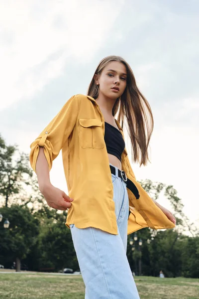 Młoda piękna kobieta w żółtej koszulce i dżinsy dreamily patrząc w aparacie stoi na trawniku w parku miejskim — Zdjęcie stockowe