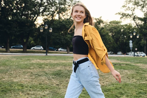Молодая красивая улыбающаяся женщина в желтой рубашке и джинсах счастливо гуляет по лужайке в городском парке — стоковое фото