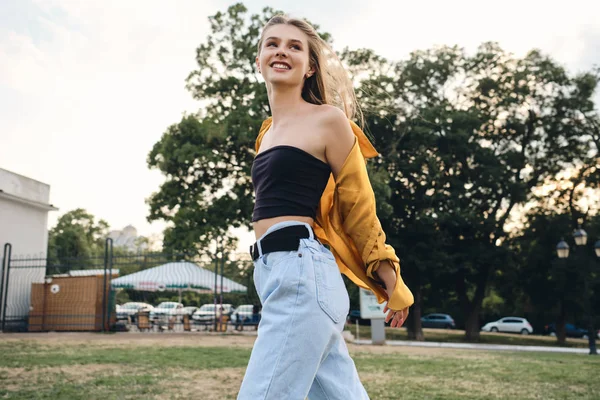 Молодая довольно веселая женщина в жёлтой рубашке и джинсах счастливо гуляет по лужайке в городском парке — стоковое фото