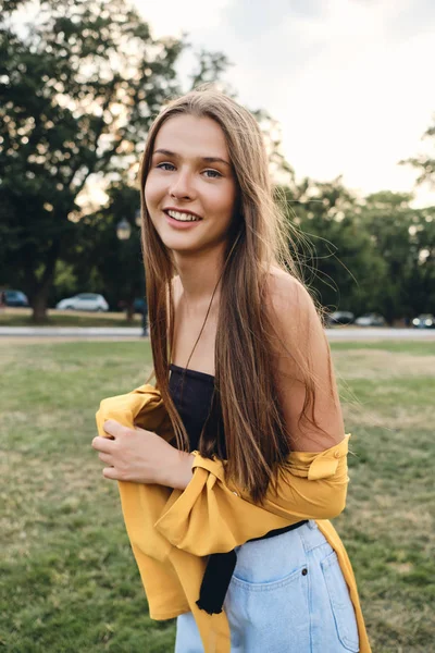 Pretty brown haired smiling girl in yellow shirt and jeans happily looking in camera standing on lawn in city park — Stock Photo, Image