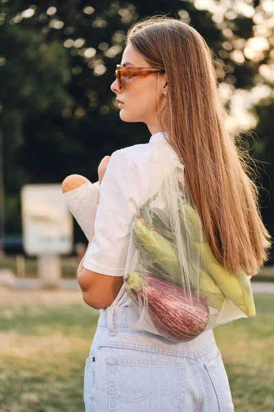 Young attractive brown haired woman standing from back with baguette bread and vegetables in eco bag on shoulder thoughtfully looking aside in city park — Stock Photo, Image