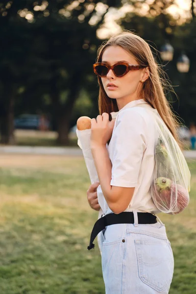 Junge hinreißende Frau in Sonnenbrille und Jeans, die Baguette-Brot und Gemüse in einer Öko-Tüte hält und nachdenklich in die Kamera im Stadtpark blickt — Stockfoto