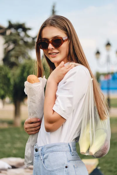 Jonge mooie lachende vrouw met Baguette brood en groenten in Eco-tas gelukkig op zoek in de camera in het stadspark — Stockfoto