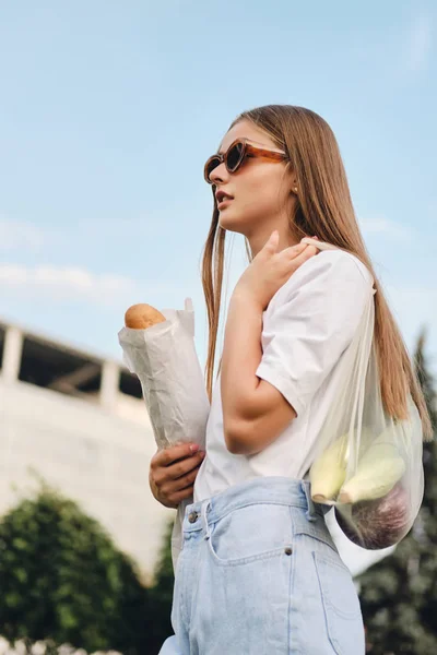 Atractiva chica de cabello castaño de pie con pan baguette y verduras en bolsa ecológica en el hombro soñando mirando a un lado en el parque de la ciudad — Foto de Stock