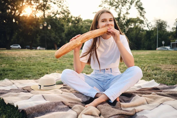 Tシャツとジーンズを着た若いかわいい女性は、チェック柄の上に座って、市内の公園でピクニックのカメラで楽しそうに見てバゲットパンを食べて — ストック写真