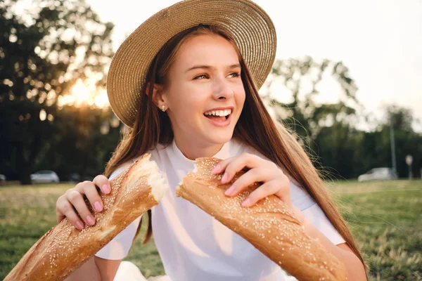 Mooie vrolijke tiener meisje in stro hoed en T-shirt gelukkig houden Baguette brood op picknick in stadspark — Stockfoto