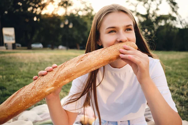 Allegro adolescente in t-shirt seduta su plaid e felicemente mangiare pane baguette sul pic-nic nel parco cittadino — Foto Stock