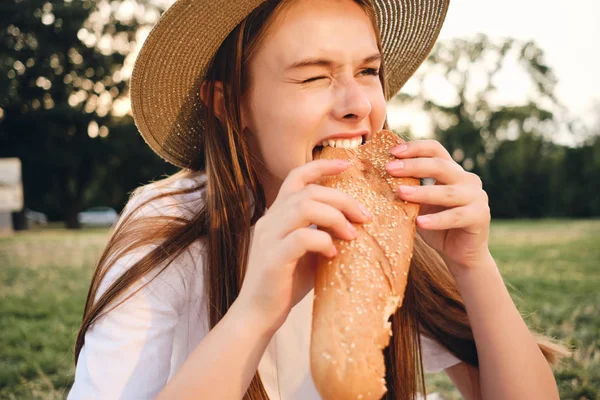 Close up attraente ragazza in cappello di paglia emotivamente mangiare pane baguette su pic-nic nel parco della città — Foto Stock