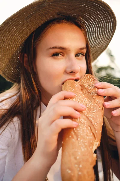 Ritratto di bella ragazza dai capelli castani in cappello di paglia mangiare pane baguette sognante in macchina fotografica su pic-nic nel parco della città — Foto Stock