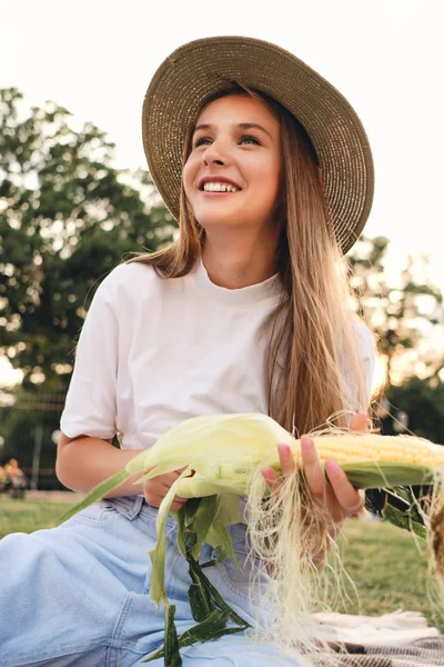 Красивая улыбающаяся каштановая девушка в соломенной шляпе радостно чистит кукурузу на пикнике в городском парке — стоковое фото