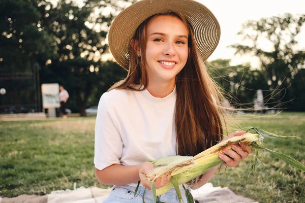 도시 공원에서 피크닉에 카메라를 찾고 행복하게 손에 옥수수를 들고 밀짚 모자에 매력적인 미소 십대 소녀 — 스톡 사진