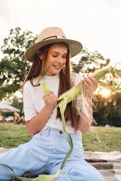 Young attractive brown haired girl in straw hat happily cleaning corn on picnic in city park — Stock Photo, Image