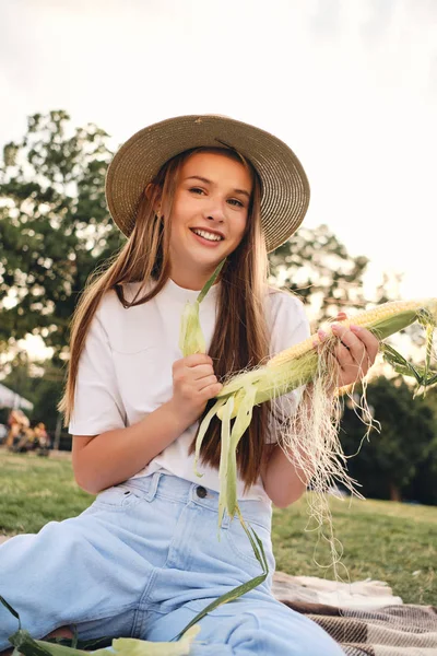 Young pretty brown haired girl in straw hat cleaning corn joyfully looking in camera on picnic in city park — Stock Photo, Image