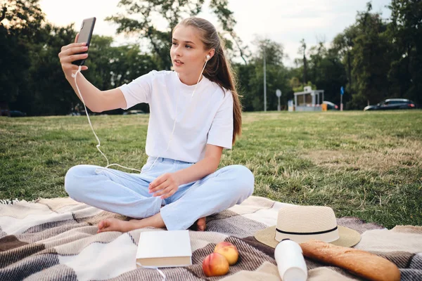 Привлекательная девушка-подросток в наушниках мечтательно фотографирует на сотовый телефон, сидя на пикнике в городском парке — стоковое фото