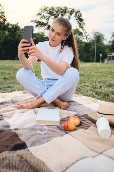 Vacker tonårs flicka i hörlurar drömmande ta foto på mobil på picknick i City Park — Stockfoto