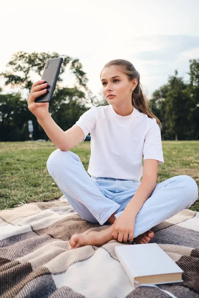 年轻迷人的女人坐在格子上看书，在城市公园的野餐中，在手机上拍照 — 图库照片