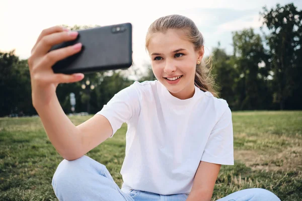Красивая улыбающаяся девушка в белой футболке счастливо фотографирует на сотовый телефон, сидя на лужайке в городском парке — стоковое фото