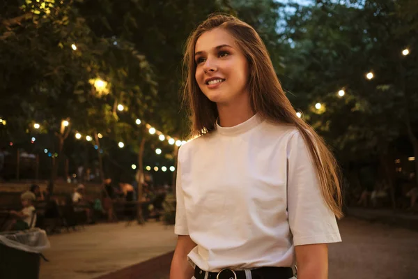 Joven bonita mujer sonriente en camiseta blanca felizmente de pie en el parque de la ciudad por la noche — Foto de Stock