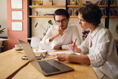 Genç iş arkadaşları düşünceli modern ofiste dizüstü bilgisayar ve kağıtları ile masada oturan yeni proje tartışıyor