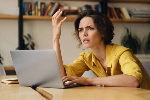 Junge brünette Frau, die empört am Tisch sitzt und im Büro am Laptop arbeitet — Stockfoto