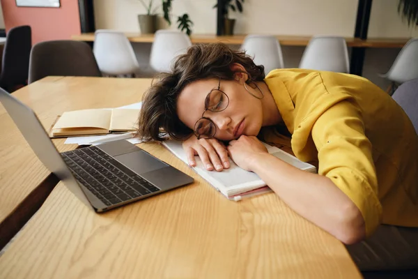 眼鏡をかけた若い疲れた女性は、職場で頭の下にノートパソコンとメモ帳を持って机の上で眠りに落ちる — ストック写真