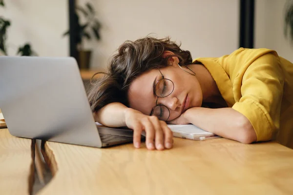 Молода втомлена жінка в окулярах спить на столі з ноутбуком на робочому місці — стокове фото