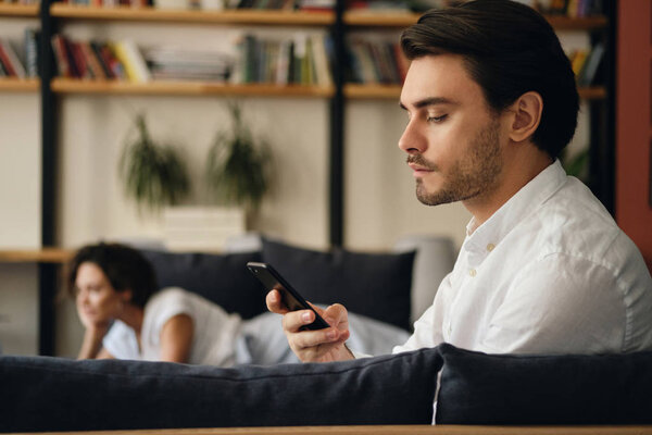 Side view of young handsome businessman sitting on sofa with colleague on background thoughtfully using cellphone in modern co-working space