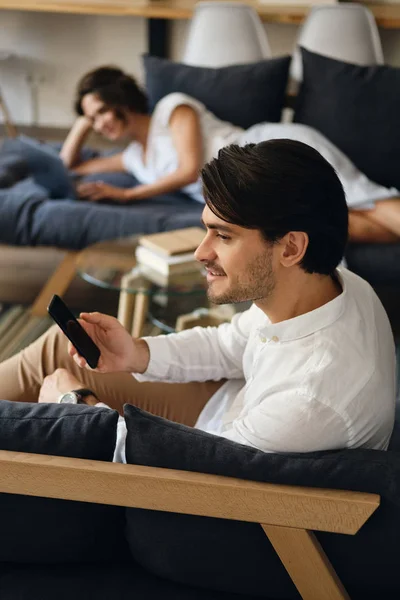 Side view of young smiling businessman sitting on sofa with cell phone happily looking aside in modern co-working space — стоковое фото