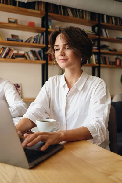 Jovem bela mulher sorridente sentada à mesa feliz trabalhando no laptop com xícara de café no escritório — Fotografia de Stock