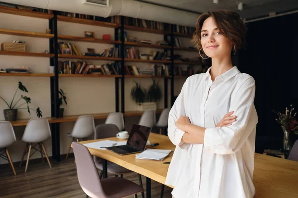 Jovem mulher atraente em camisa branca sonhadoramente olhando na câmera com mesa no fundo no escritório moderno — Fotografia de Stock