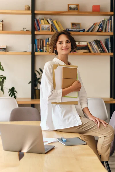 Mulher sorridente atraente nova na camisa branca que olha feliz na câmera que se senta na mesa com papéis e portátil no escritório moderno — Fotografia de Stock