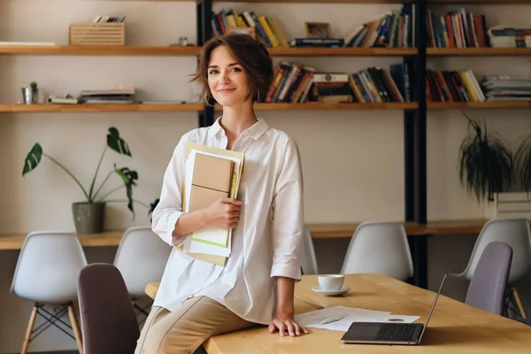 Молодая привлекательная женщина в белой рубашке счастливо смотрит в камеру, сидя на столе с бумагами и ноутбуком в современном офисе — стоковое фото