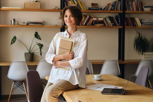Beyaz gömlekli genç güzel kadın mutlu modern ofiste kağıtları ve dizüstü bilgisayar ile masada oturan kamera bakıyor — Stok fotoğraf